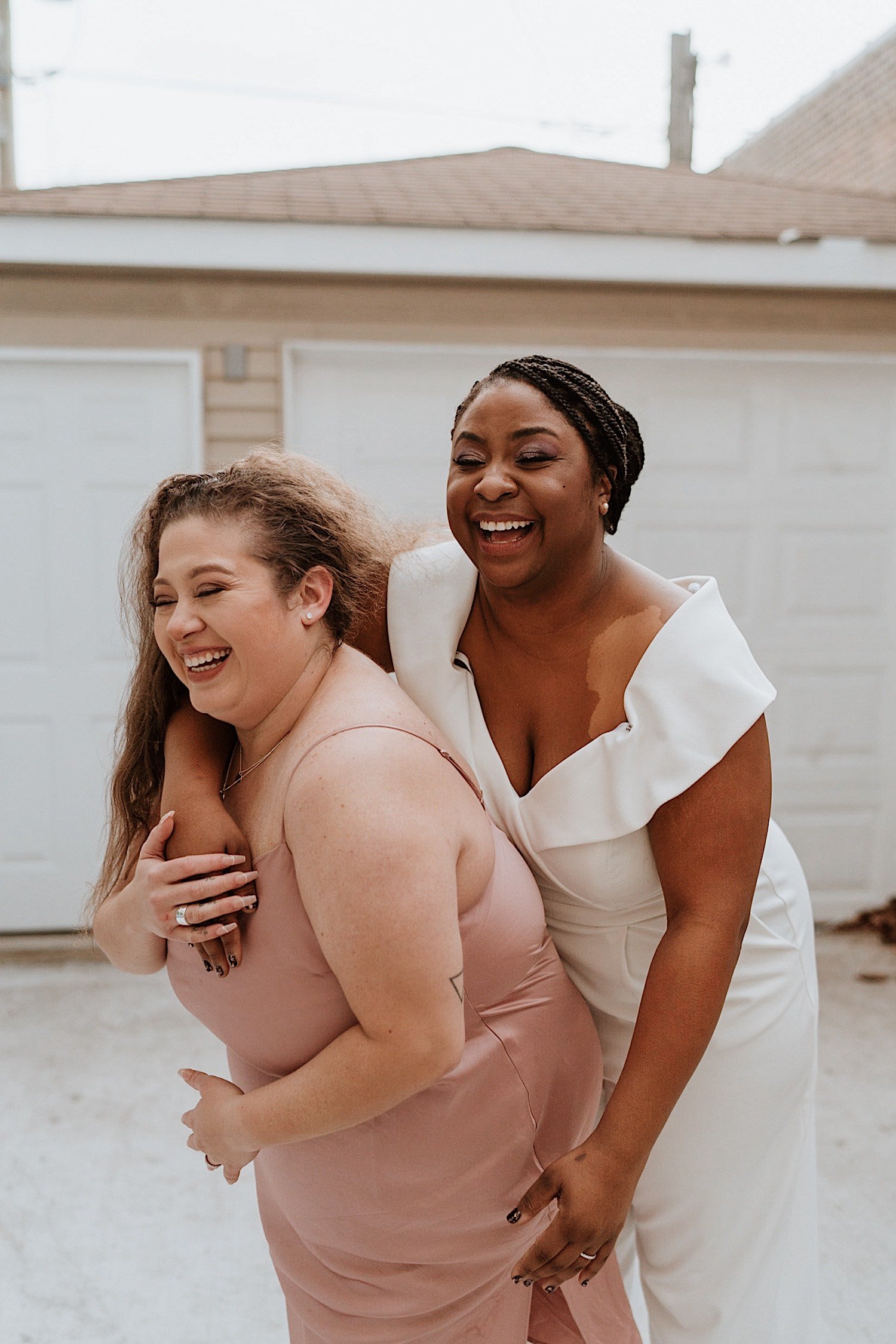 Brides laugh and hold each other in front of garage