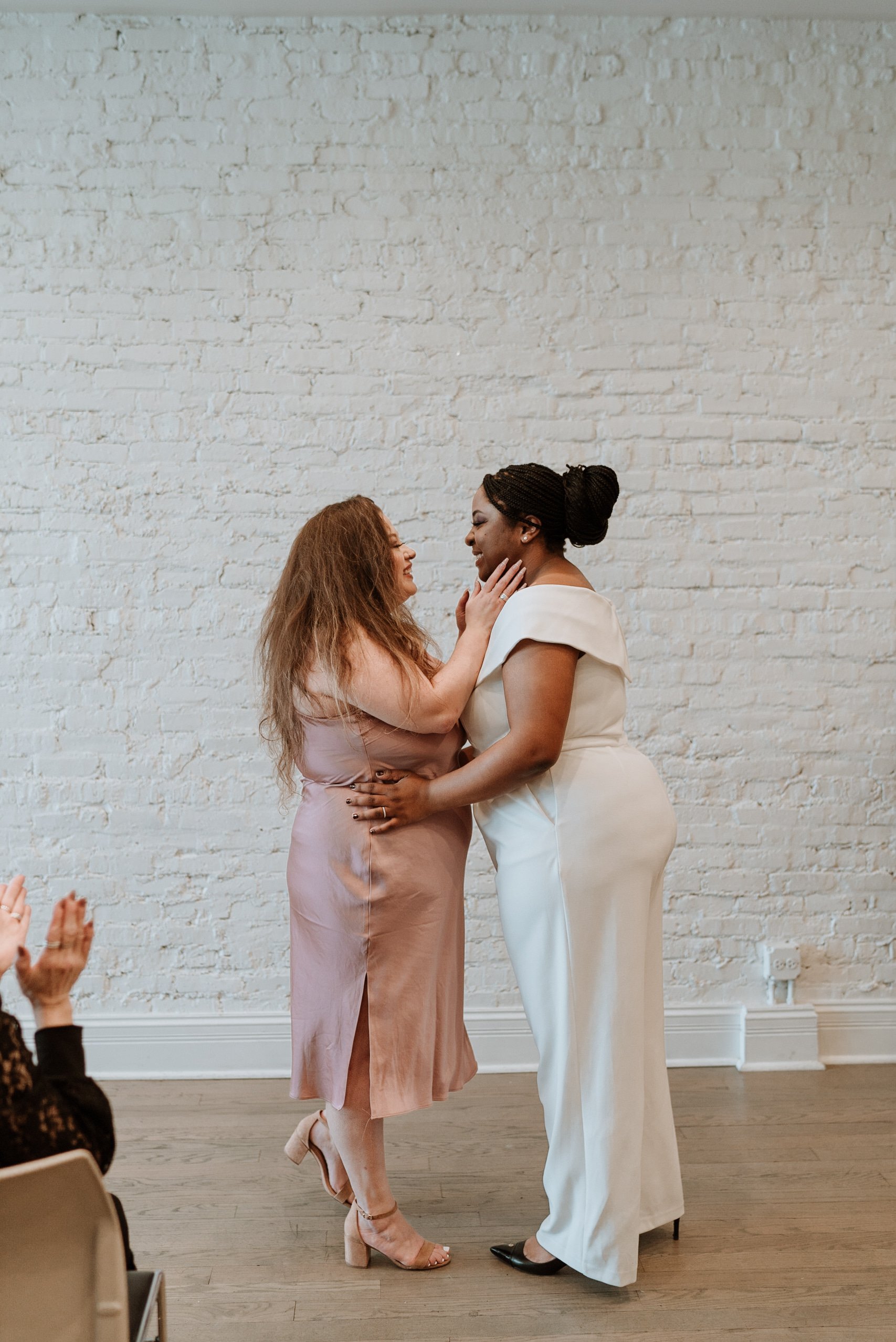 Brides embrace each other after their wedding ceremony in front of white wall of their Humboldt Park venue