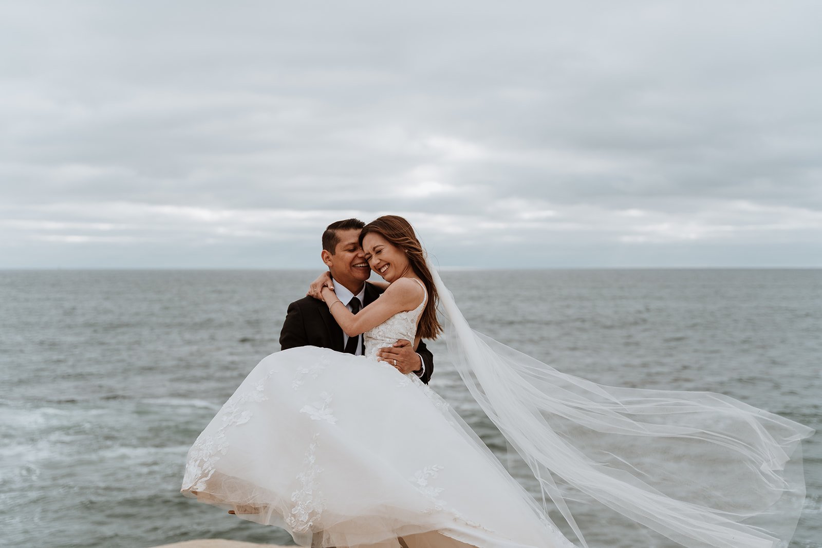 Groom holds bride while in wedding gear as they laugh standing in front of the ocean