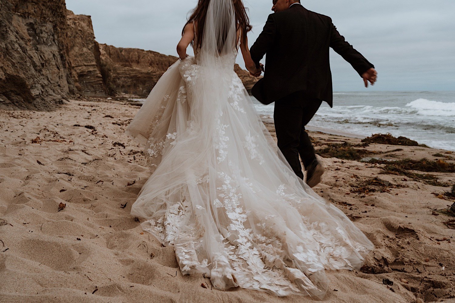 Bride and groom wearing wedding gear run on the beach in front of the water