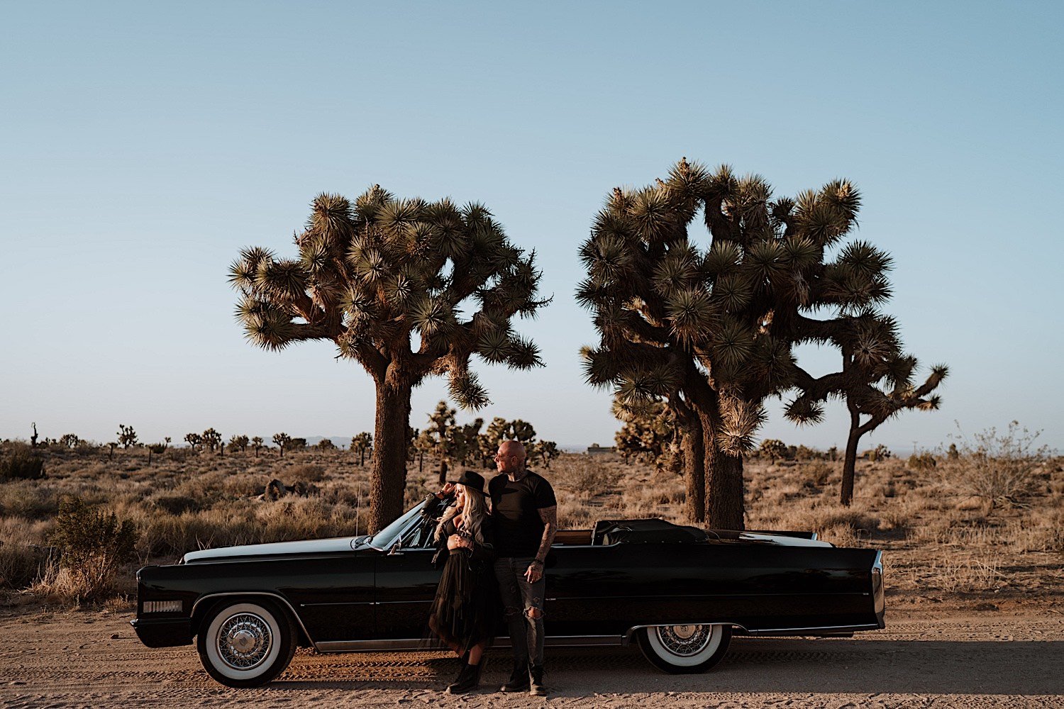 Couple posed next to a classic car in the desert in front of Joshua trees