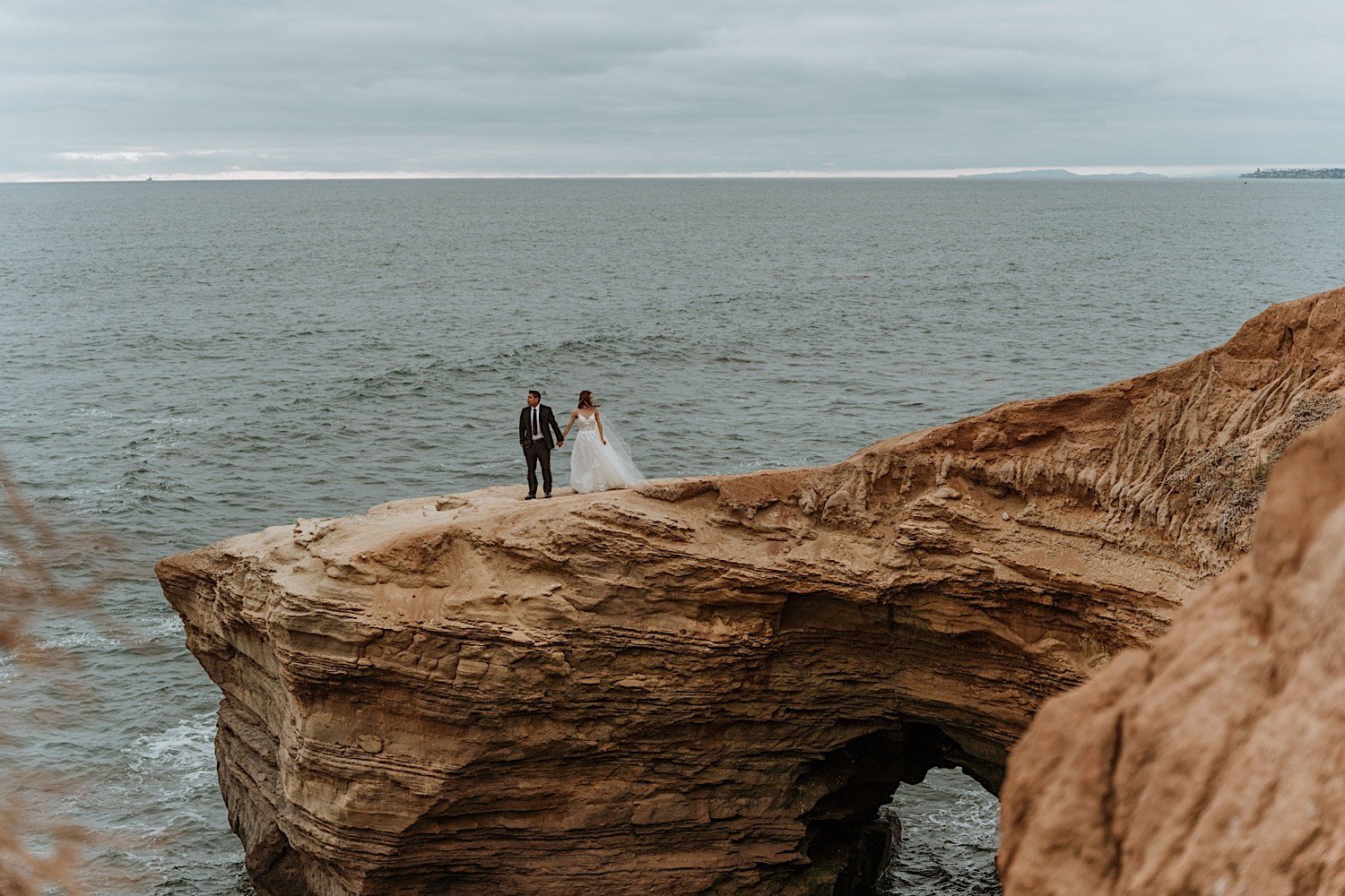 Bride and groom hold hands and look opposite directions while standing on a cliff overlooking the ocean