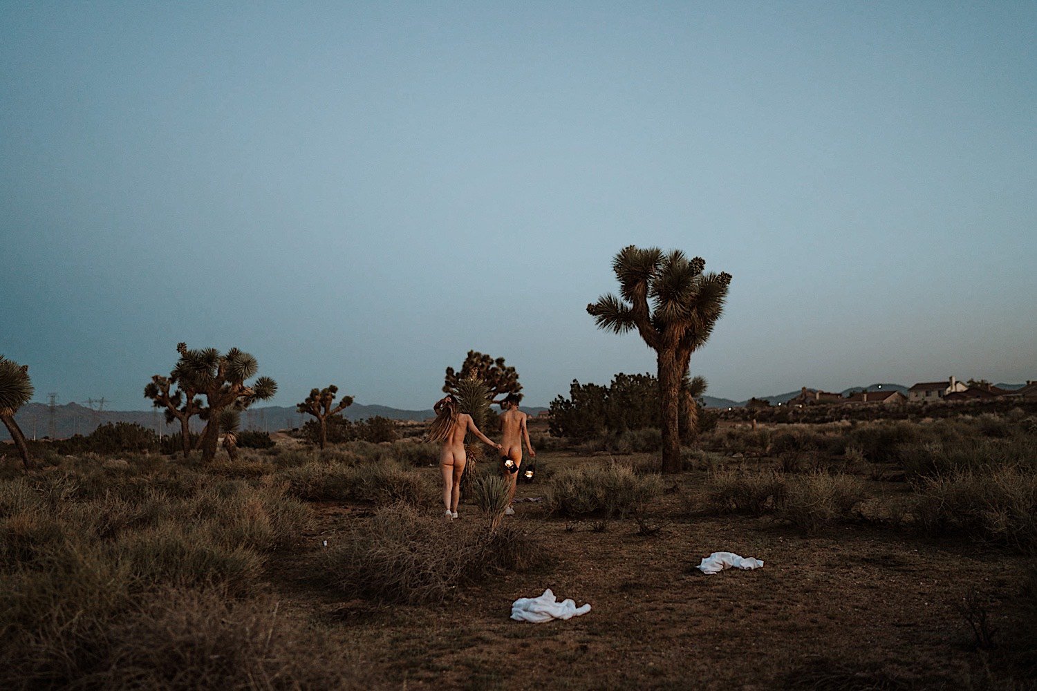 Naked couple walk away from the camera holding lanterns in the desert with Joshua trees around them