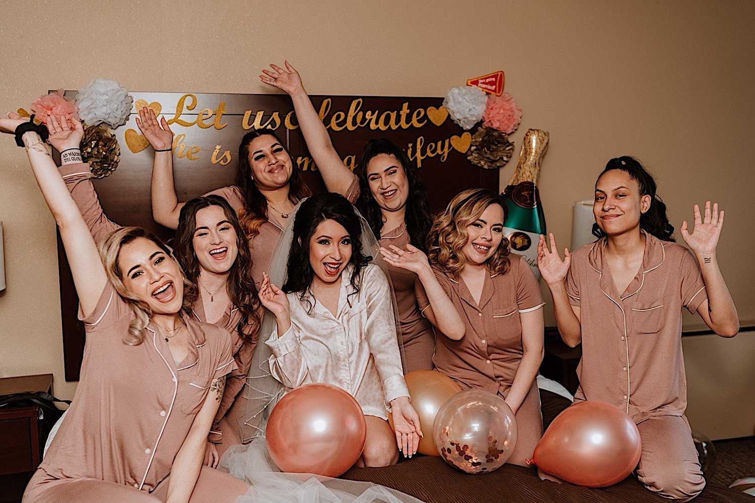 Bride and bridesmaids sit in bed and wave to the camera with balloons