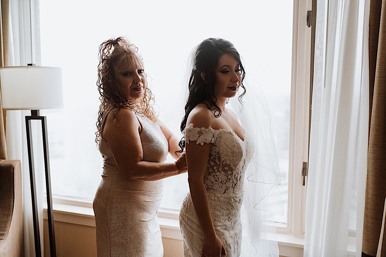 Mother helps bride zip up the back of her wedding dress in front of a window