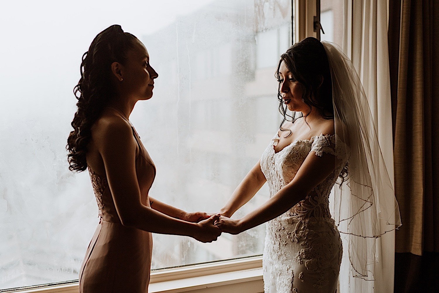 Bride and bridesmaid hold hands in front of a window before wedding