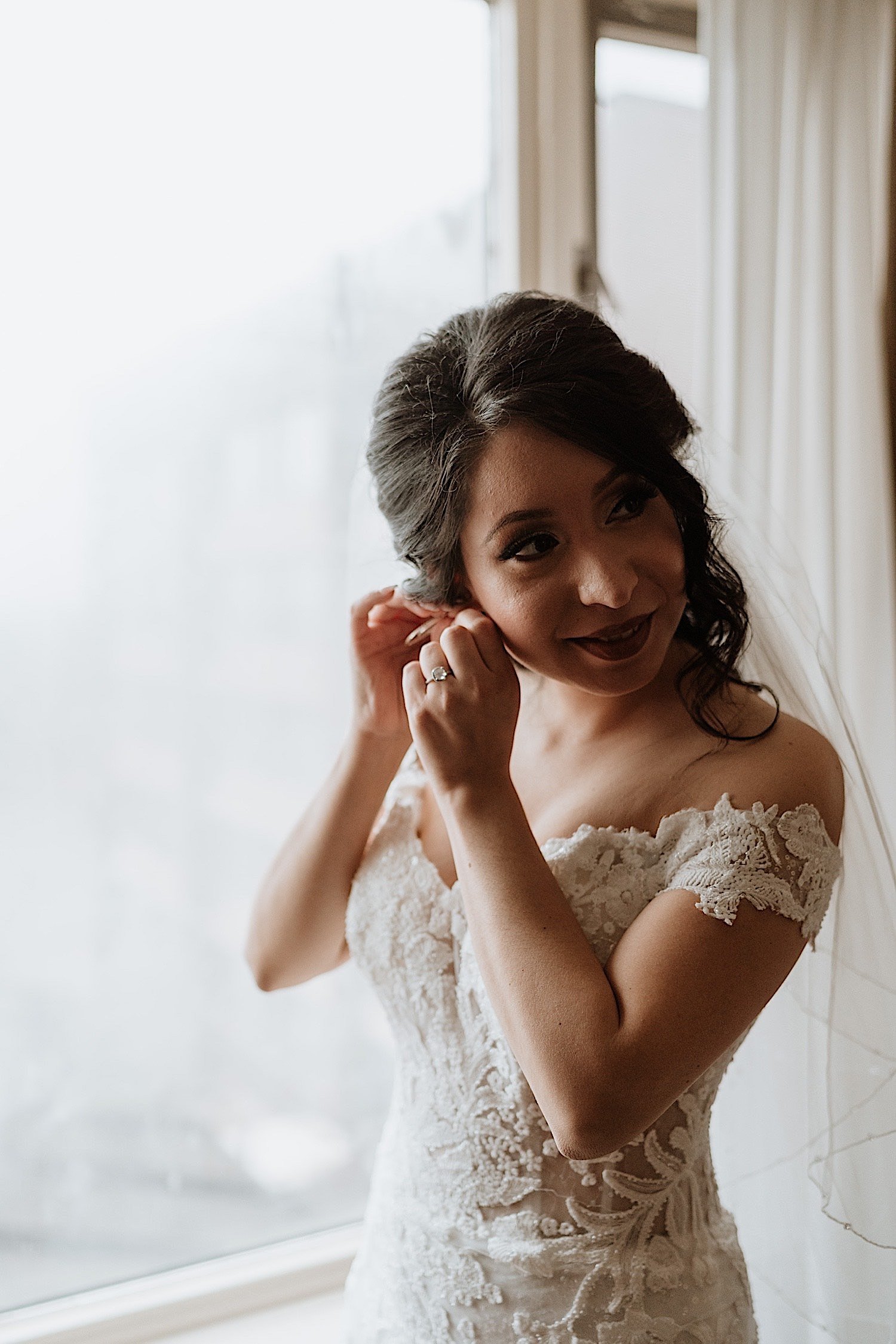 Bride putting on an earing while in her wedding dress in front of a window