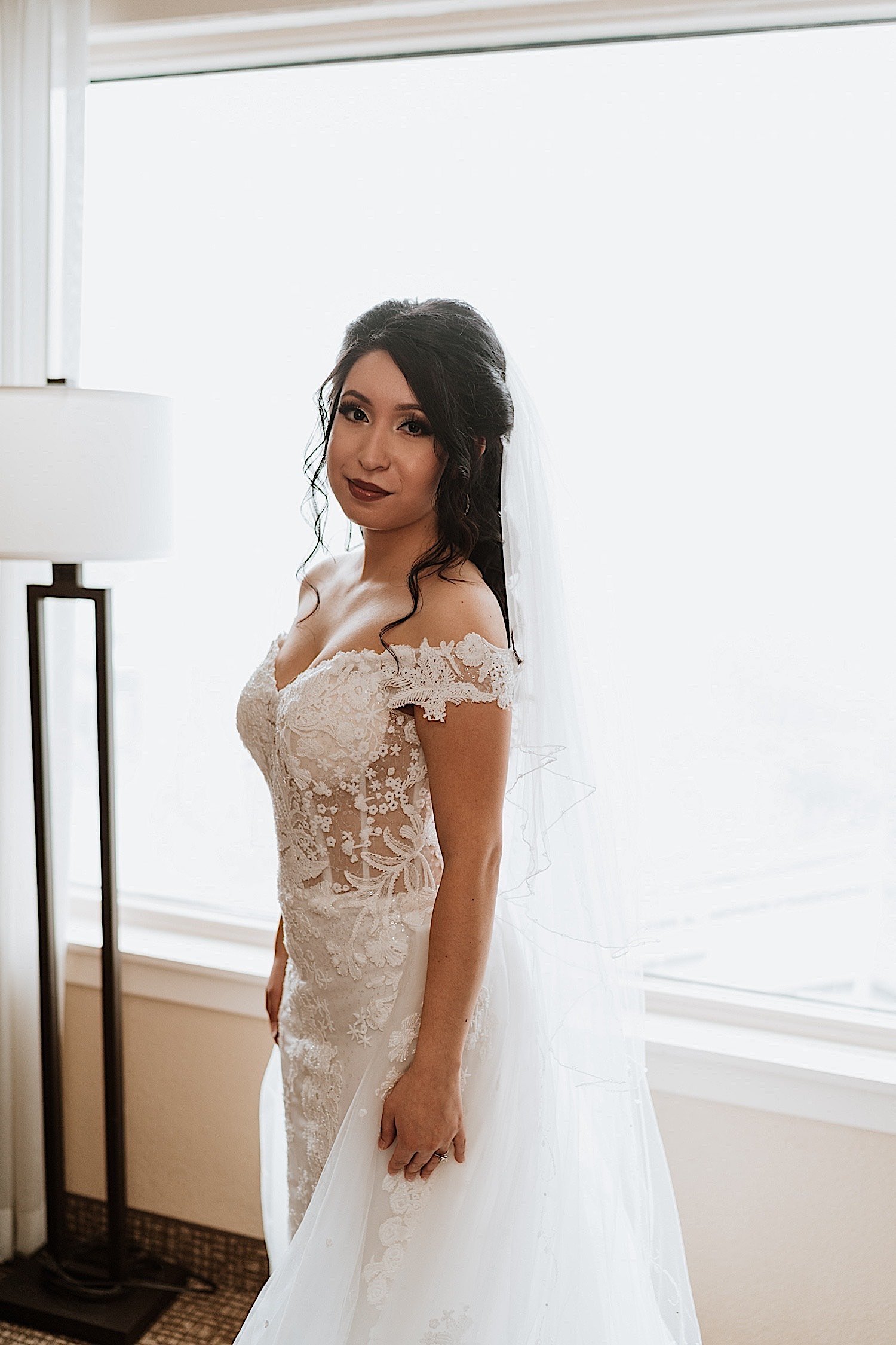 Bride poses in her wedding dress and looks at the camera in front of a window