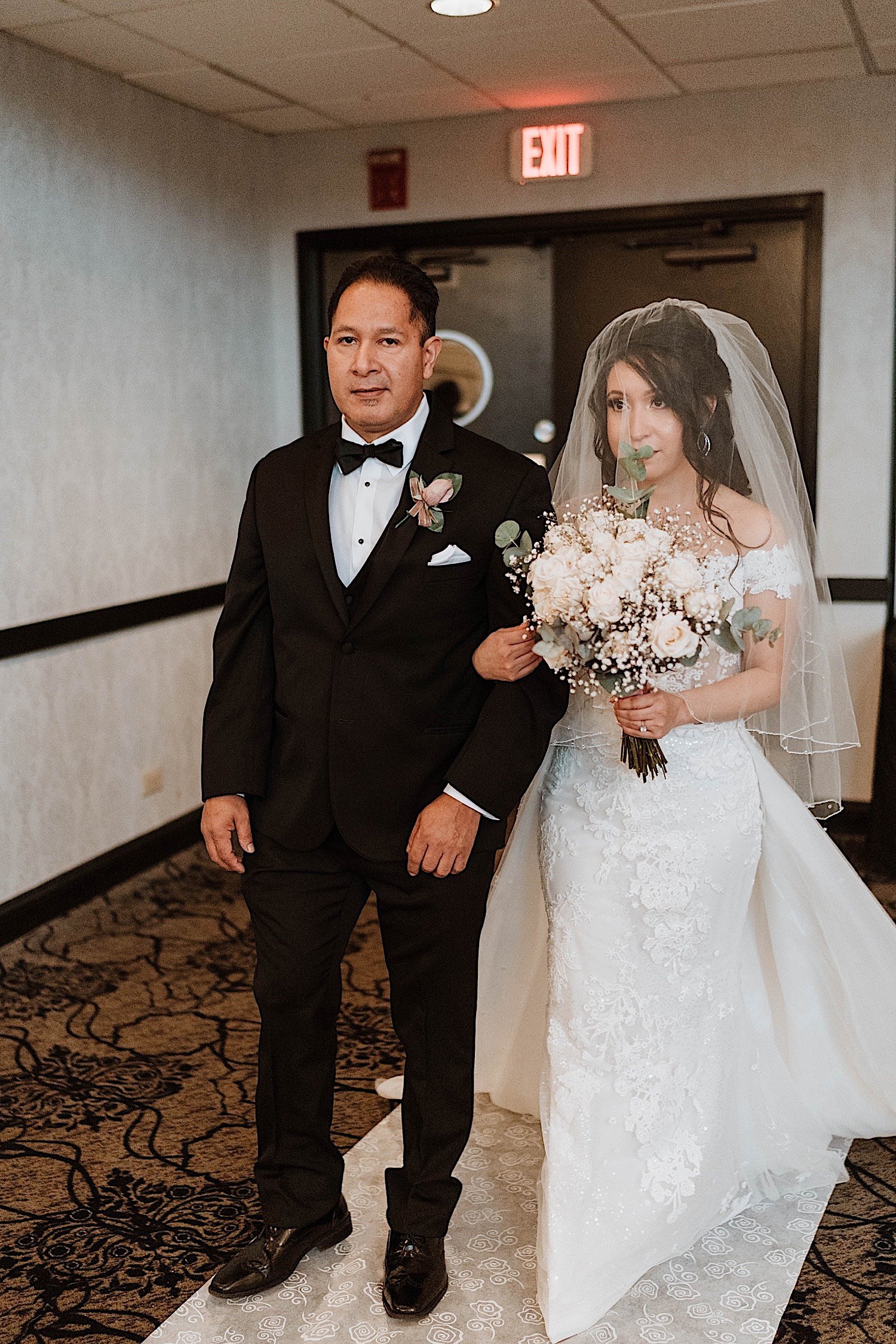 Bride enters her ballroom wedding ceremony escorted by her father while holding a bouquet of flowers