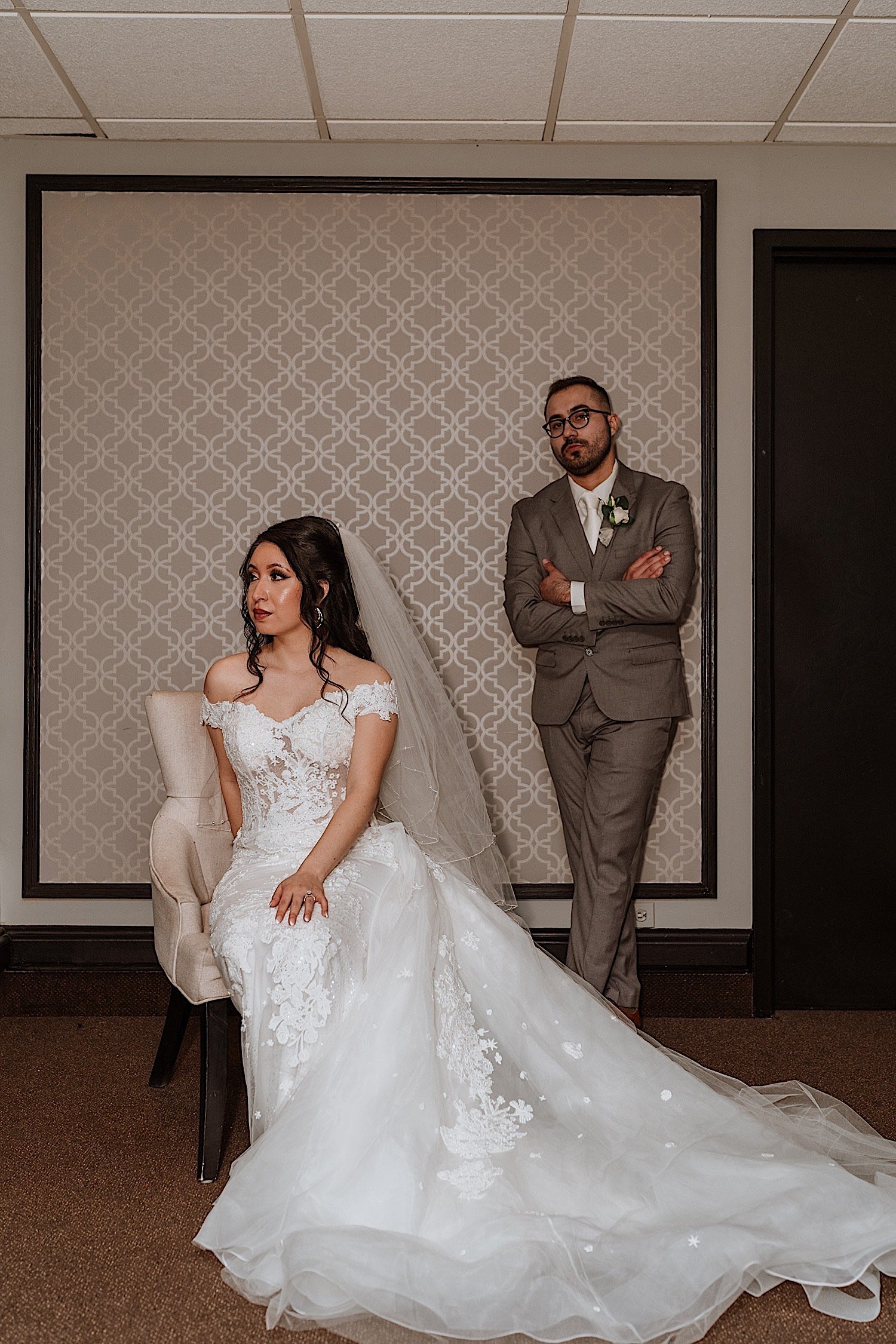 Groom stands behind seated bride as she looks away while he looks at the camera
