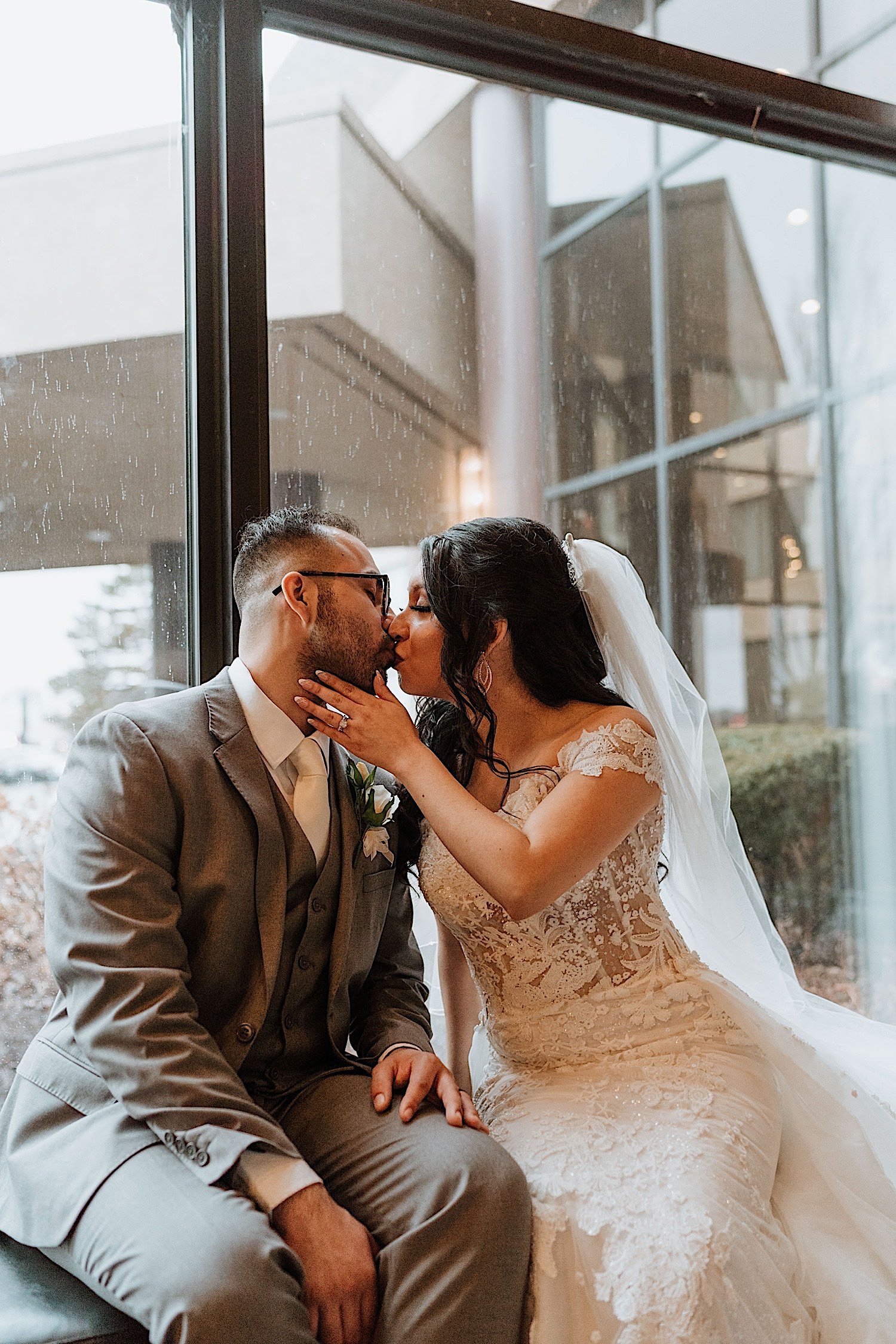 Bride and groom kiss while seated in front of a window while it rains outside