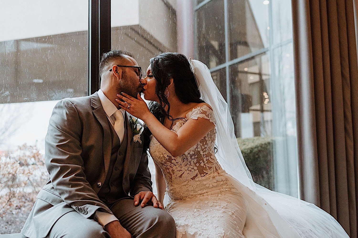 Bride and groom kiss while seated in front of a window as it rains after their Chicagoland ballroom wedding