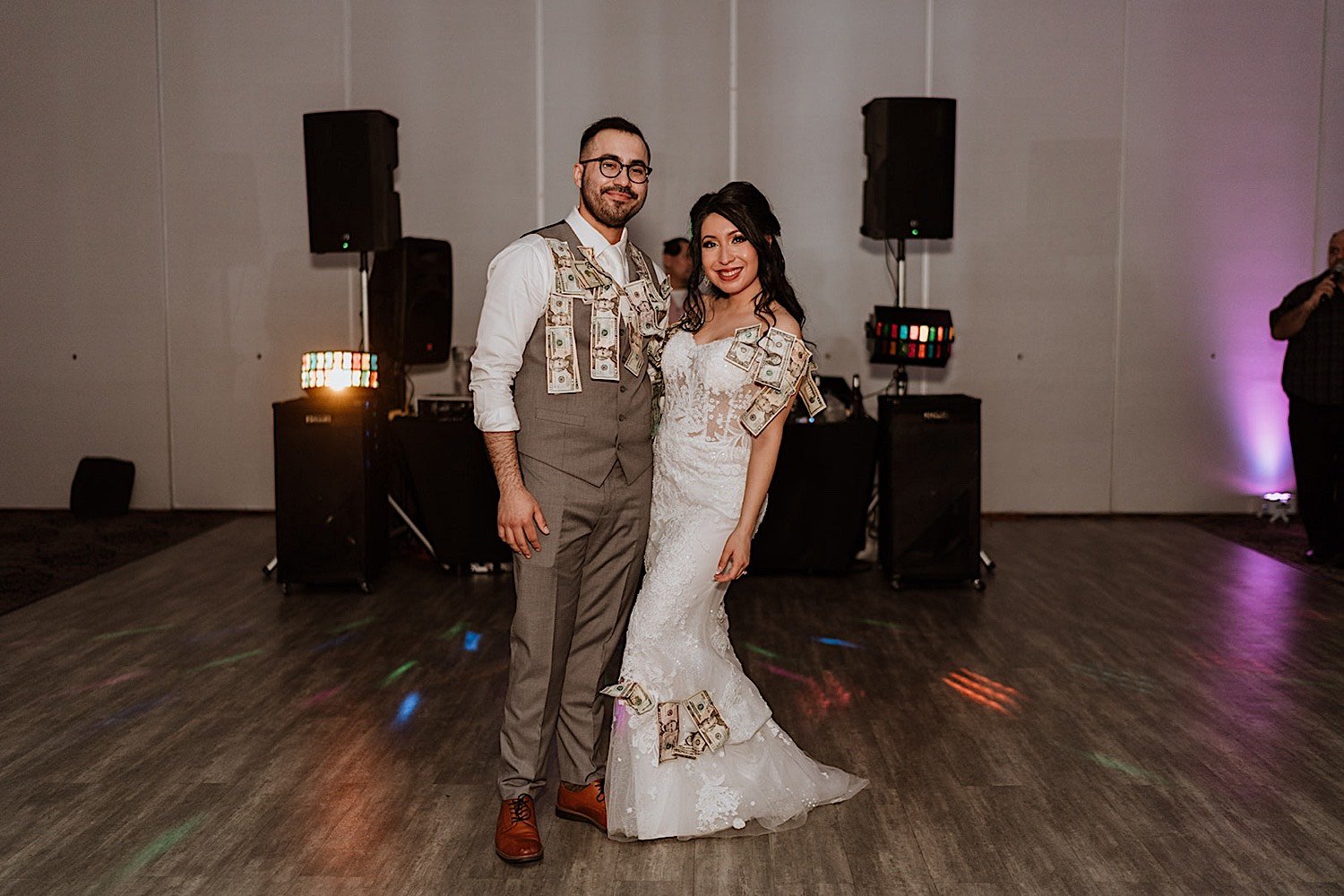 Bride and groom pose on dance floor with money pinned to them in front of DJ