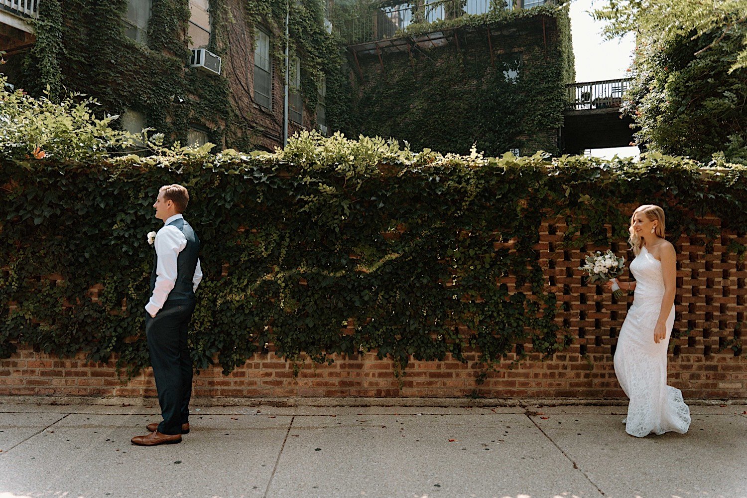 Bride walking up behind groom to surprise him with a first look in front of a vine covered brick wall