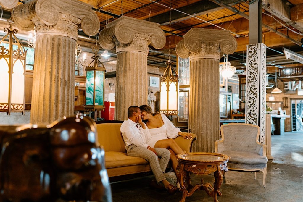 A couple smiles and almost kisses while sitting on a vintage couch surrounded by grecian pillars and vintage chandeliers at Salvage One in Chicago Illinois.