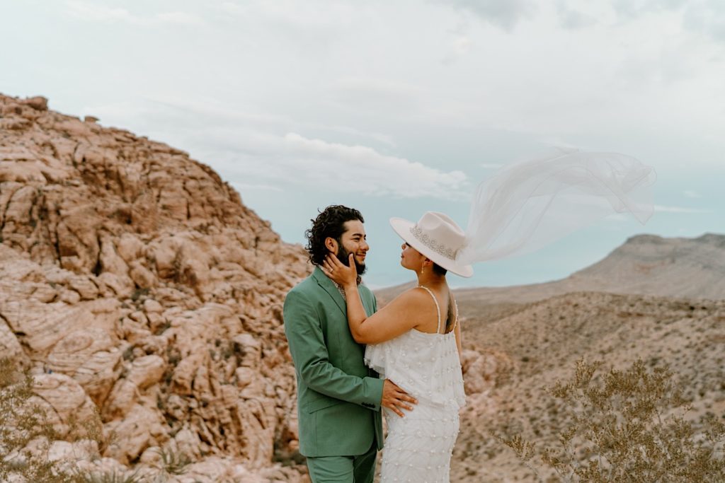 A bride and groom hold one another in front of the Red Rocks, the bride is wearing a two piece white dress and wide brimmed hat with a veil.  The groom his wearing a green suit.