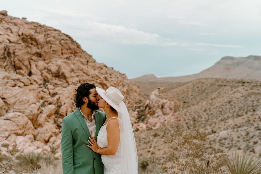 A bride and groom kiss in front of the Red Rocks, the bride is wearing a two piece white dress and wide brimmed hat with a veil.  The groom his wearing a green suit.
