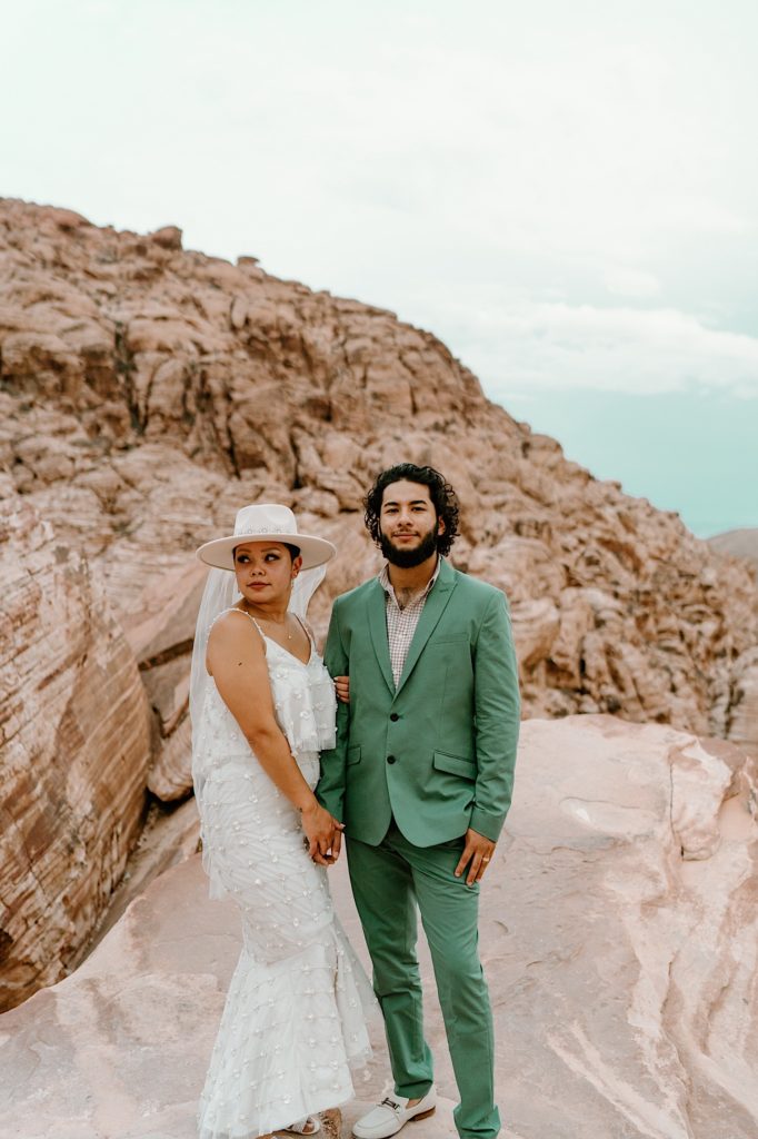 A bride and groom hold one another in front of the Red Rocks, the bride is wearing a two piece white dress and wide brimmed hat with a veil.  The groom his wearing a green suit.
