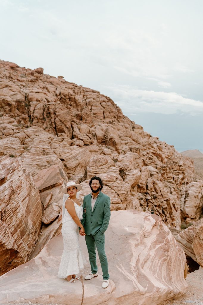 A bride and groom stand holding hands in front of the Red Rocks, the bride is wearing a two piece white dress and wide brimmed hat with a veil.  The groom his wearing a green suit.