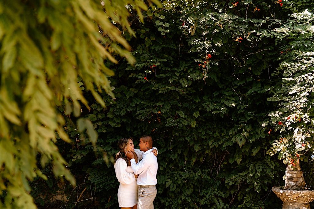 A couple snuggles in close to one another while smiling and holding each other.  They are standing in front of a brick wall covered in Ivy.