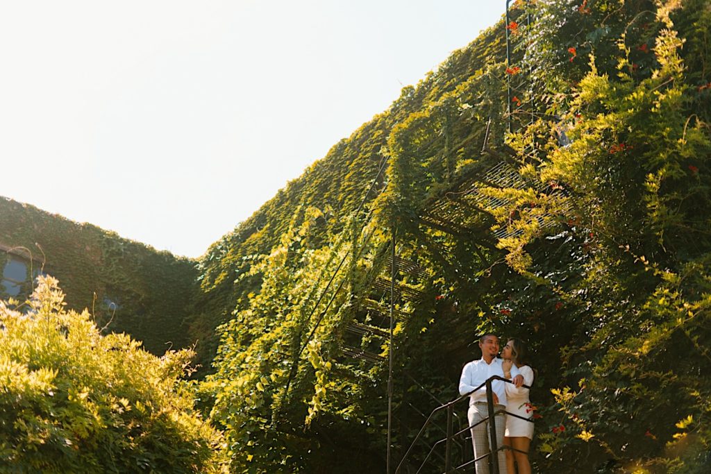 A couple smiles at one another while standing on a metal fire escape at Salvage One wedding venue.  The fire escape is covered in Ivy so they stand out in a see of green leaves.
