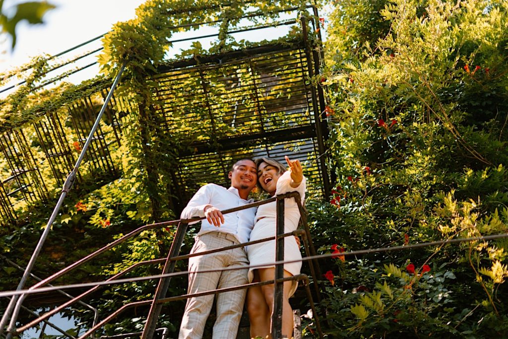 A couple stands on a metal stairwell covered with moss at Salvage One in Chicago.  The couple is showing off their engagement ring and smiling directly at the camera.