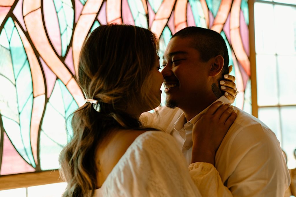 A couple stands in front of pink, purple and green stained glass smiling and almost kissing.  