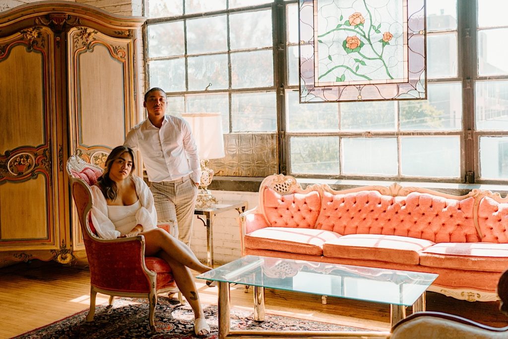 A couple looks directly at the camera while sitting in a room with a brass vintage table and pink vintage couches during their engagement session.