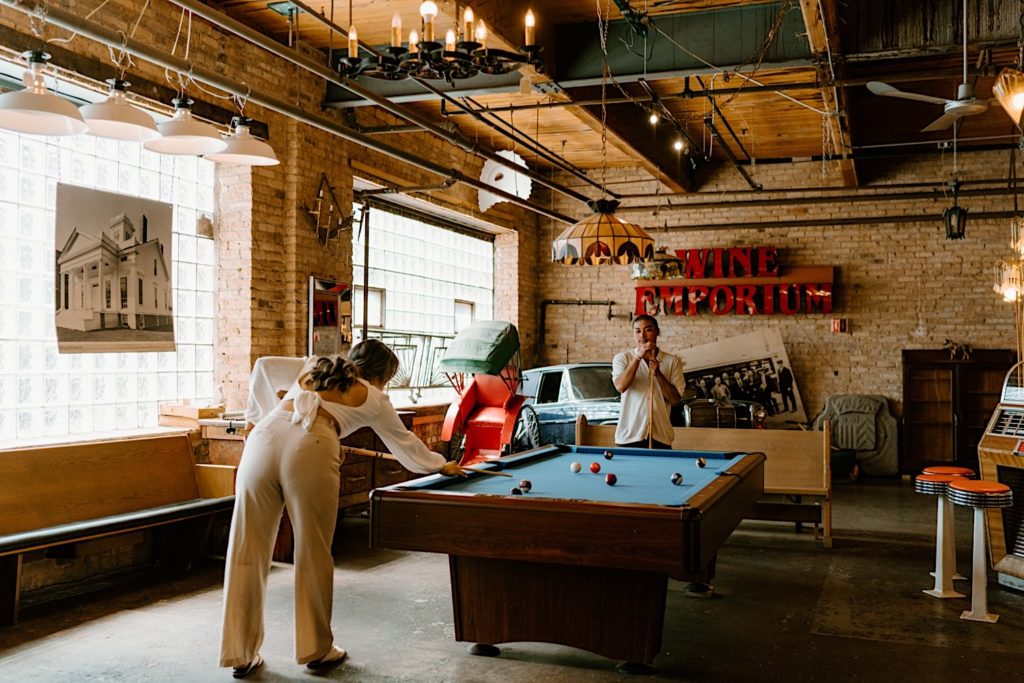 A couple plays pool during their Chicago engagement session at Salvage one, they are leaning over the pool table deciding their next moves.  There is a giant sign that says wine emporium behind them along with a vintage car.