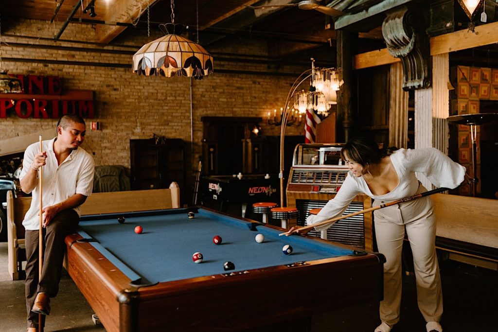 A couple plays pool on a pool table with blue felt during their engagement session at Salvage One.  There is a vintage juke box in the background and a variety of vintage lights.