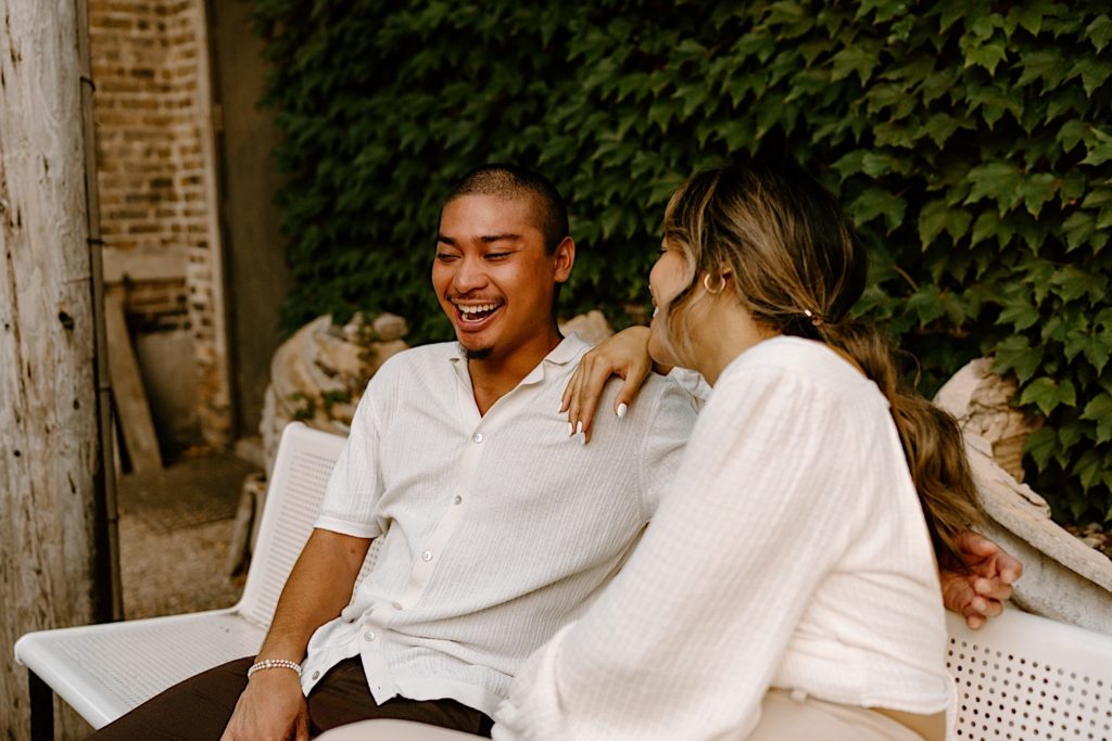 An engaged couple sits outside on a white metal bench laughing.  They are taking their engagement session in the courtyard at Salvage One, a Chicago wedding venue.