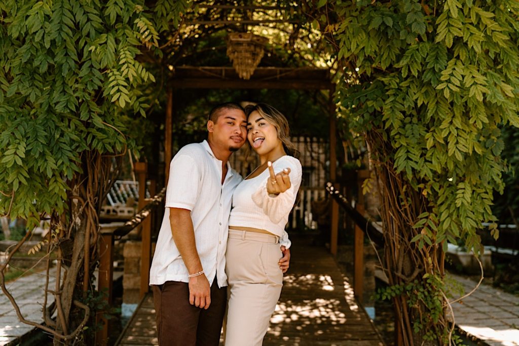 A fiancée shows off her ring while sticking her tongue out and holding her fiancé close.  They are standing under a pergola covered in leaves at Chicago's Salvage One.