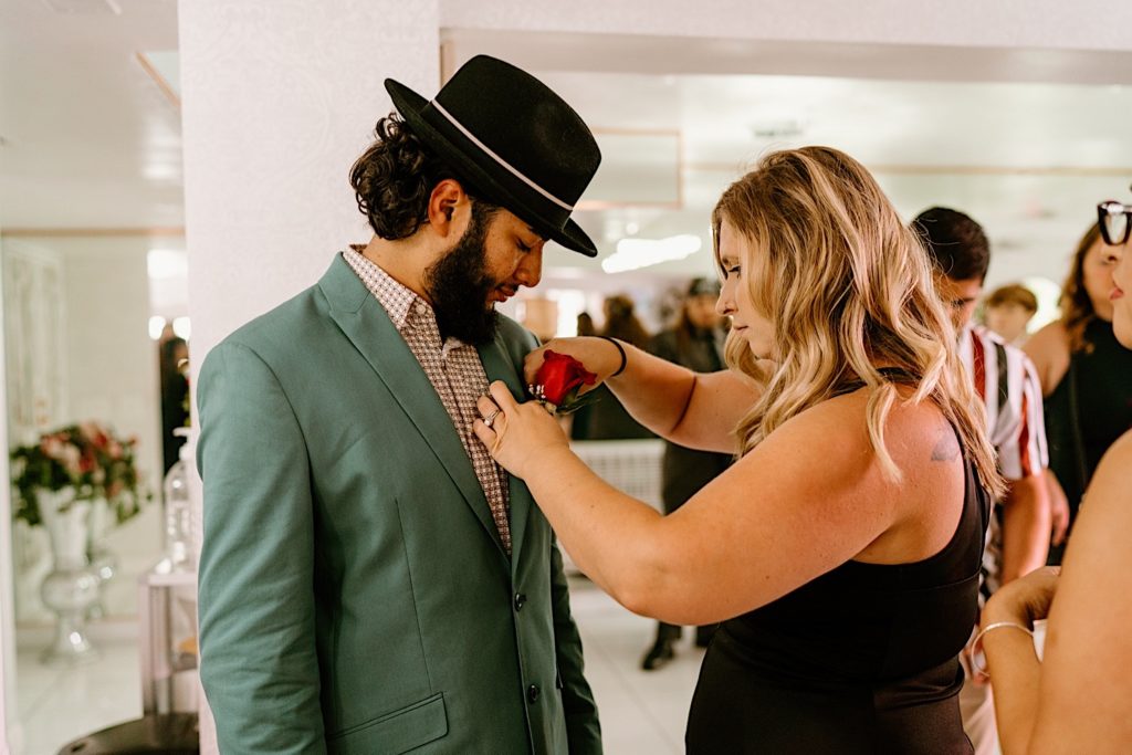 A friend of the groom pins a boutonniere on the grooms green suit coat.