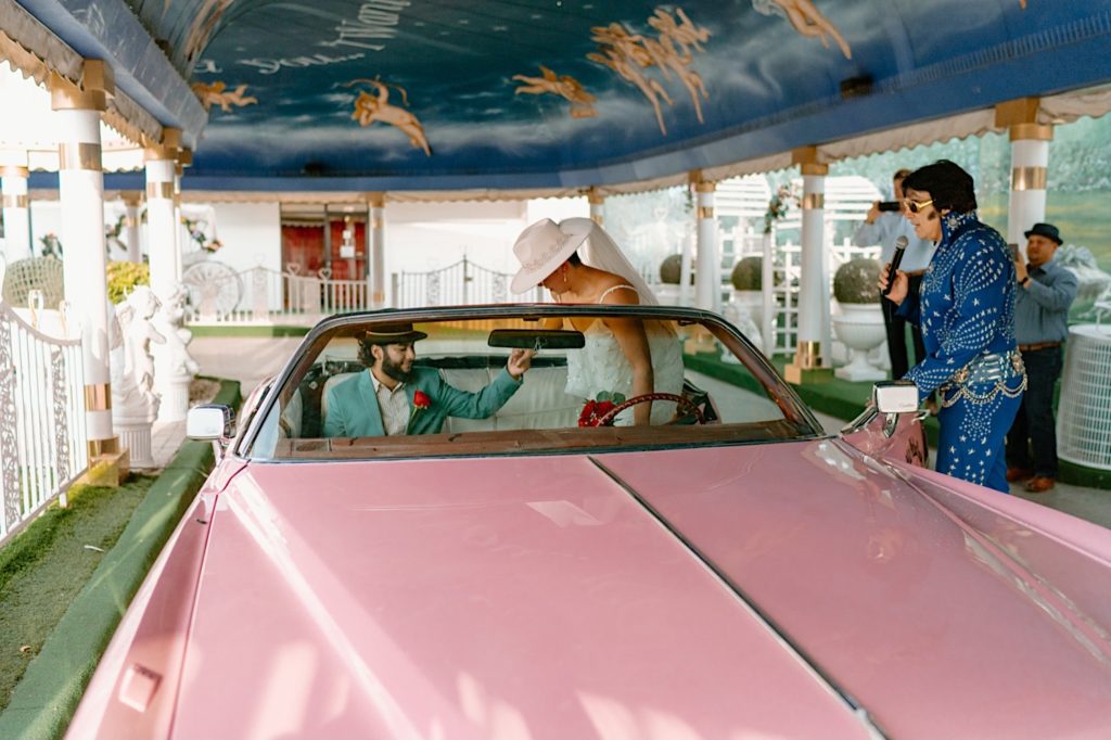 The groom holds his brides hand as they climb into a pink convertible so that they can be married by an Elvis impersonator.