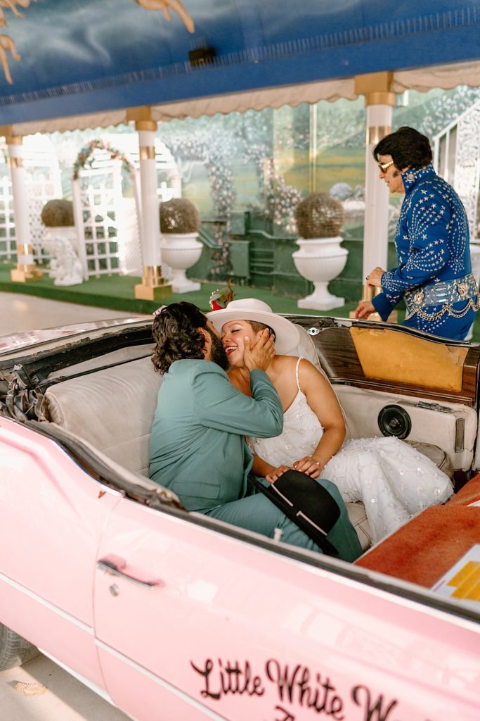 The bride and groom kiss in the pink convertible while Elvis oversees the last of their wedding ceremony.