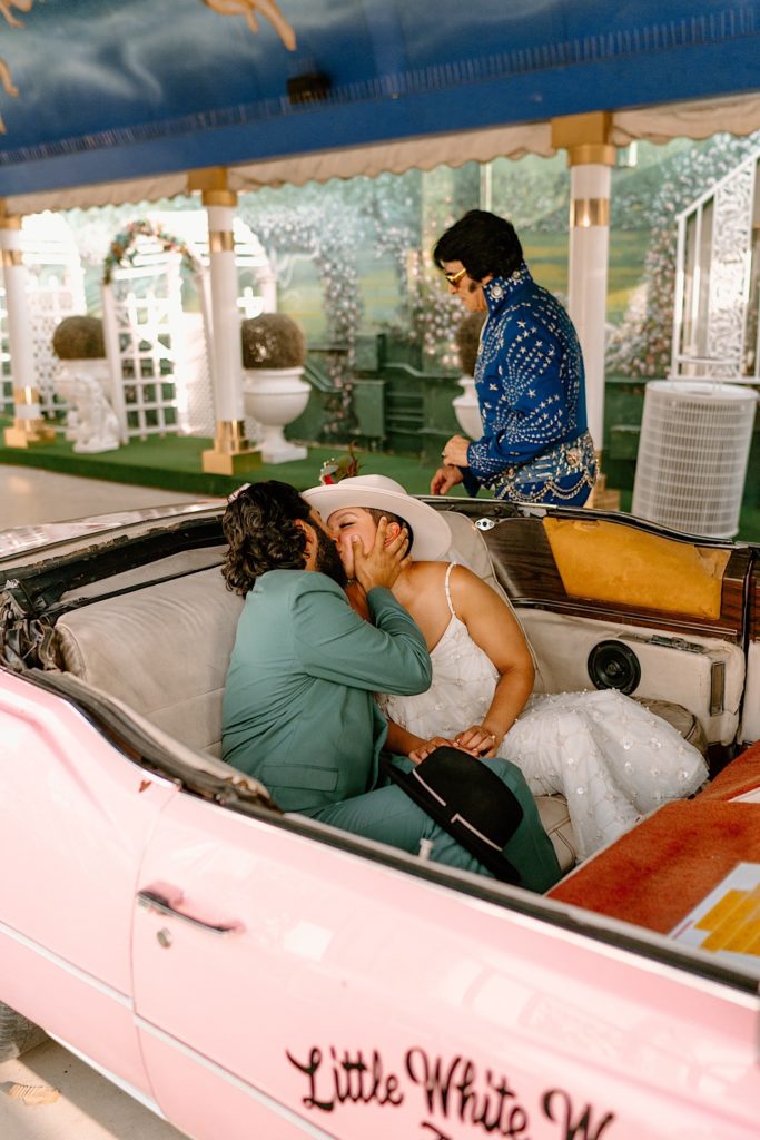 The bride and groom kiss in the pink convertible while Elvis oversees the last of their wedding ceremony.