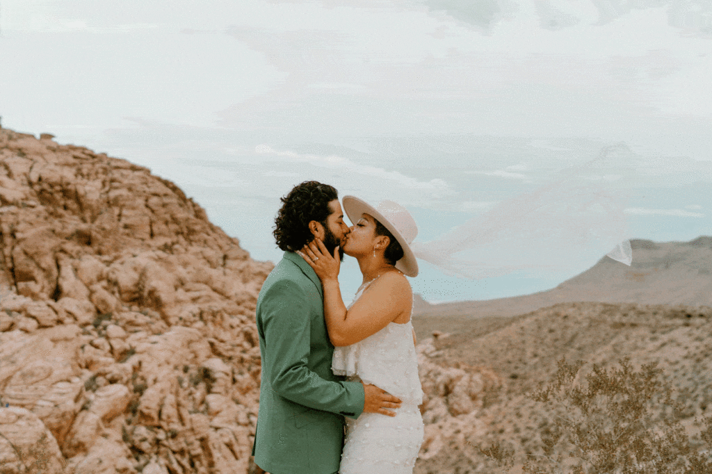 A bride and groom kiss in front of the Red Rocks, the bride is wearing a two piece white dress and wide brimmed hat with a veil.  The groom his wearing a green suit.
