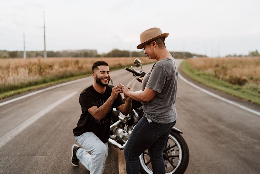 A fiancée cries as her boyfriend proposes to his girlfriend next to his motor cycle on a spring day.
