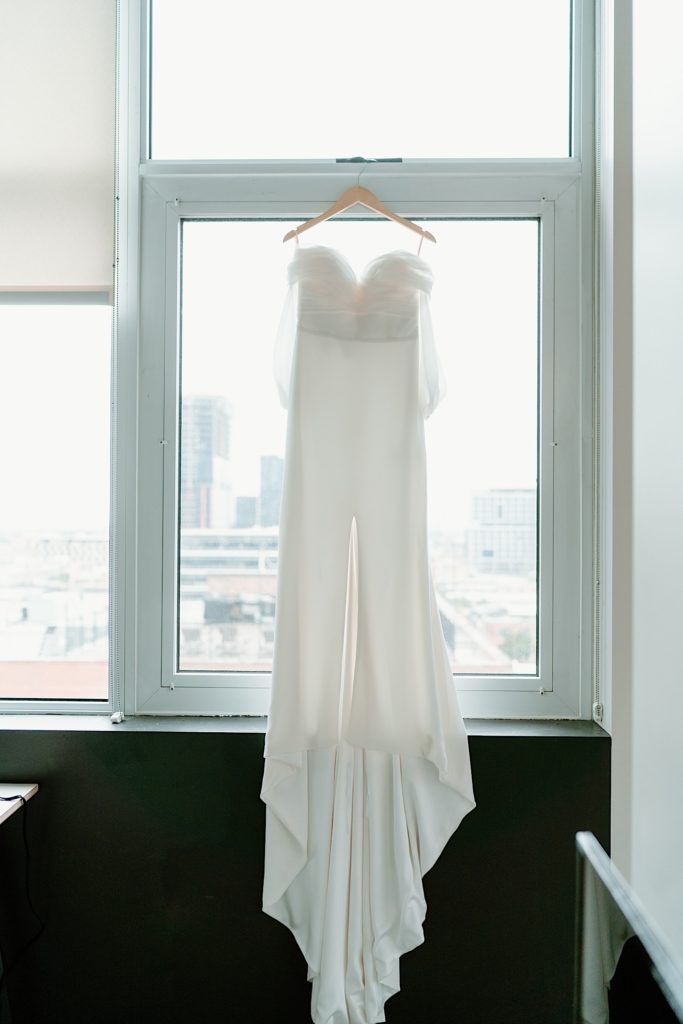 A white wedding dress hanging from a window in the getting ready room.