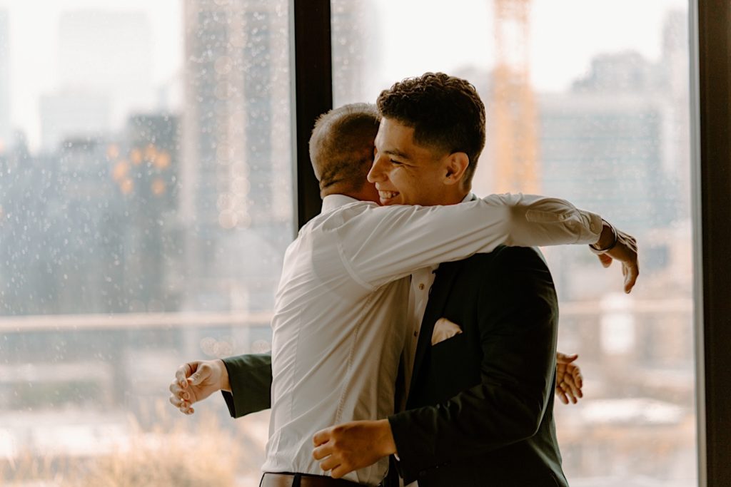 The groom is helped into his jacket by his father overlooking the City of Chicago from a window behind them.  