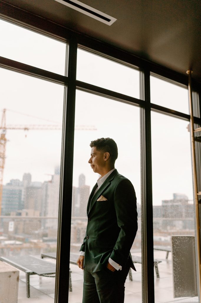 The groom stands with his green suit in front of a window on his wedding day in his Chicago Airbnb