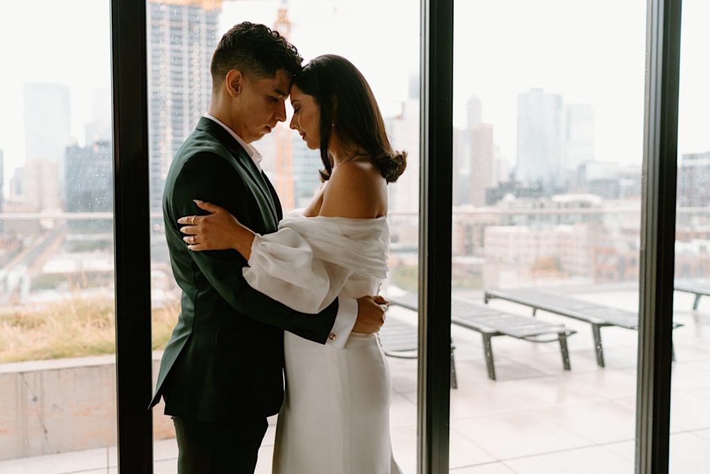 A bride and groom hold one another in front of a window overlooking the city before their wedding at Loft Lucia.