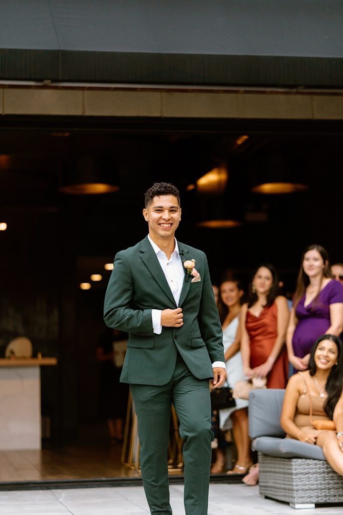 The groom walking into his ceremony at Loft Lucia wearing a green suit and with a pink boutonnière.