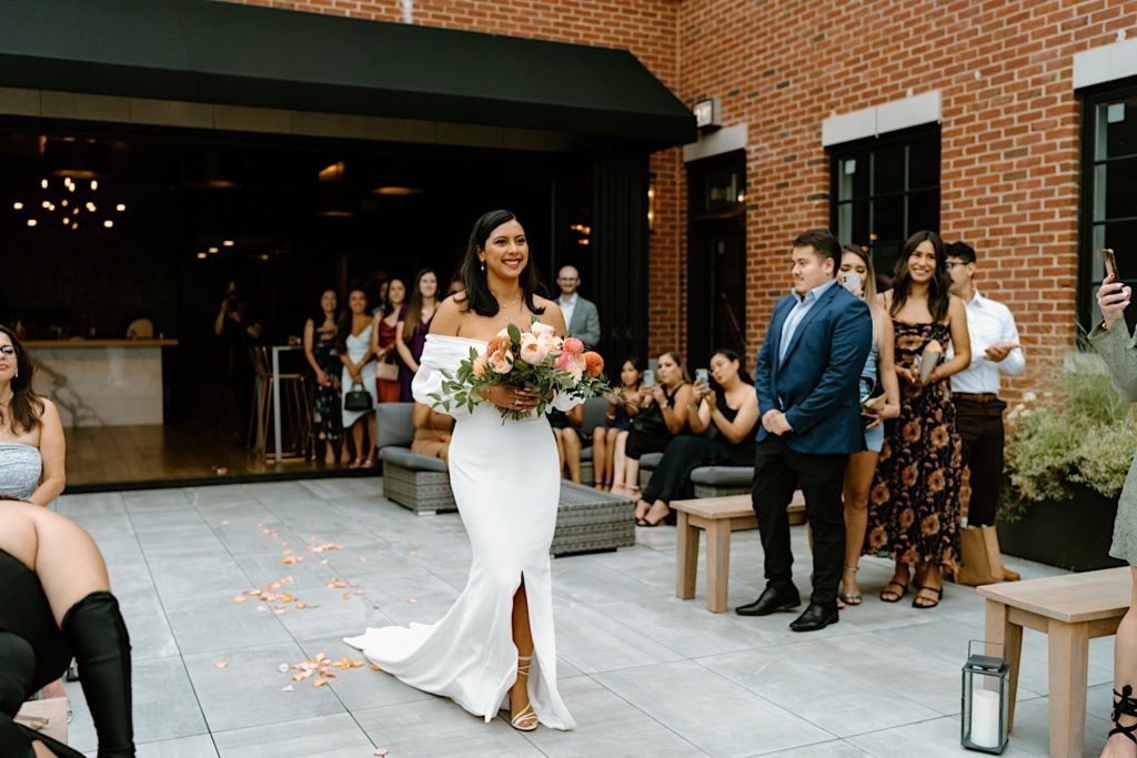 The bride walks down the aisle with a pink bouquet towards her groom in the ceremony space at Loft Lucia.