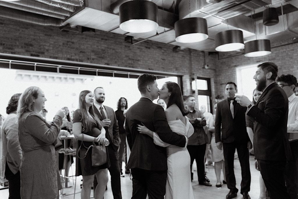 The bride and groom kiss surrounded by their friends and family celebrating in their reception space in Chicago.