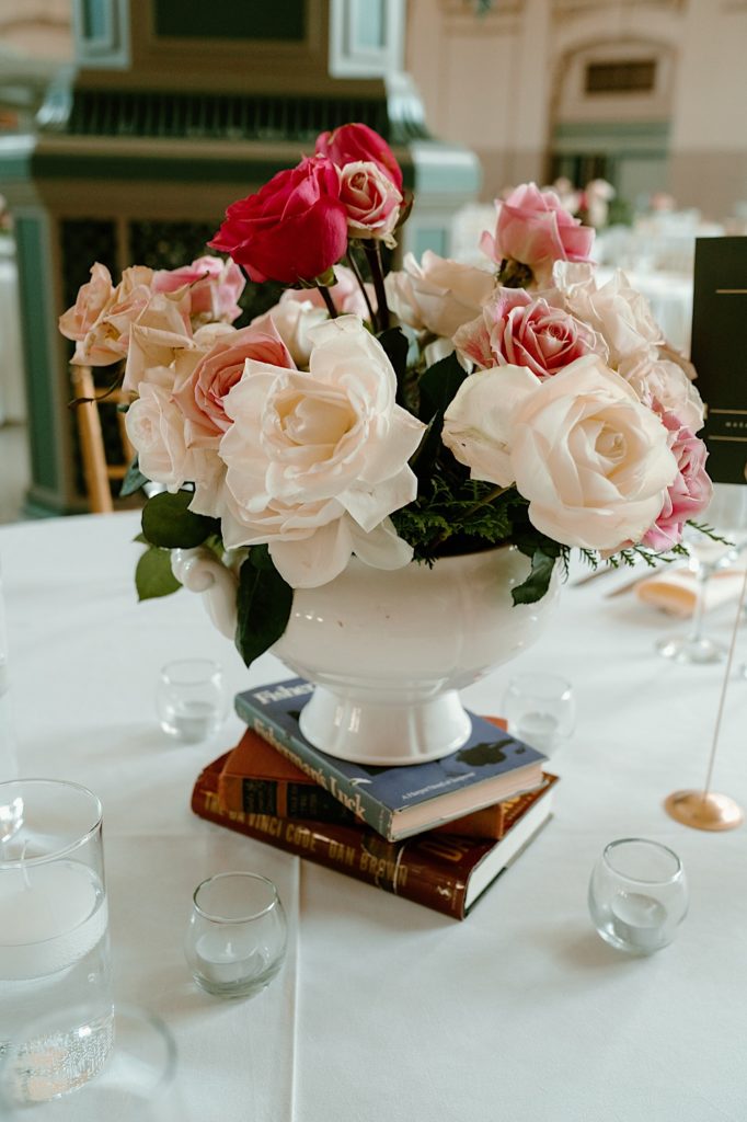 A wedding centerpiece with a white vase, pink flowers and books.
