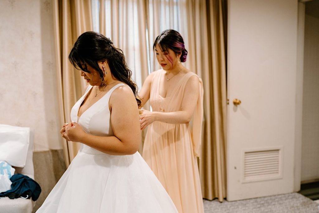 A bride stands getting ready for her wedding with one of her bridesmaids buttoning the back of her dress before their ceremony at Joliet's Union Station.