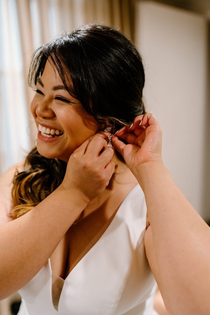 A bride putting in her teardrop earrings and smiling before her wedding ceremony.
