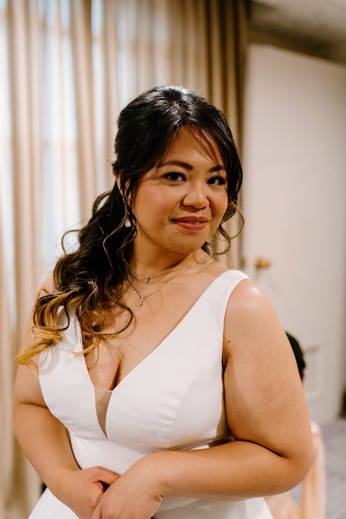 A bride smiling at the camera before with a white wedding dress with a low v neckline.