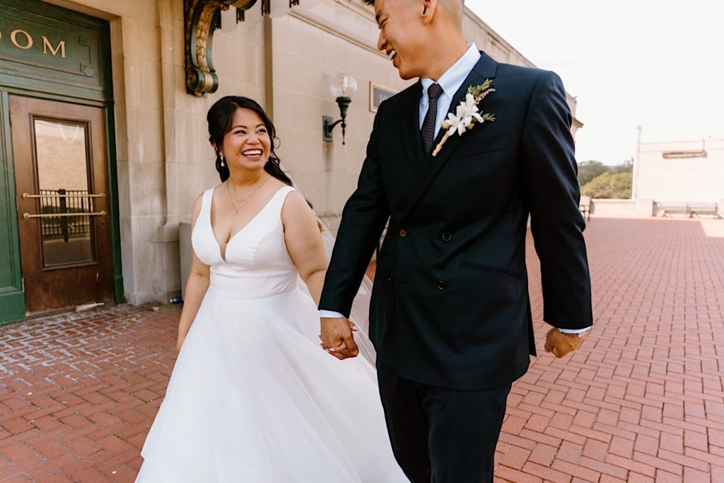 A bride and groom hold hands walking towards the camera smiling at one another at A bride and groom stand holding one another during their first look standing underneath the overhand at Joliet's Union Station.